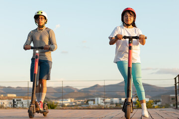 Latino tourists riding electric scooters in the city at dusk, wearing safety helmets and...