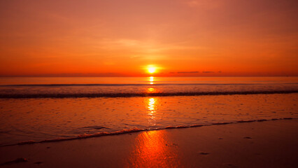 Fototapeta premium Beautiful red sunrise on the beach. The sun rises above the horizon and calm sea waves on the sand in the foreground. Romantic meditative nature summer landscape