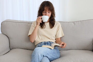 Beautiful curly brunette drinking coffee and reading a book on the couch at home