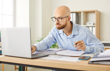 Bald man in glasses sitting at desk in bright office, focused on his work. Busy company employee looking at laptop screen while holding pen with documents and files spread across desk in front of him.