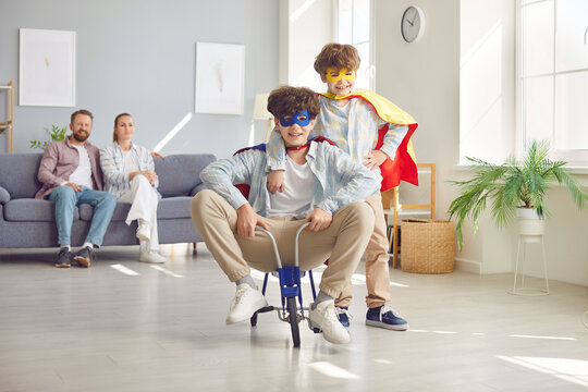 Happy playful family, parents and excited funny little brothers at home wearing super hero fancy costume riding small tricycle, carefree two generations family having fun in new spacious living room