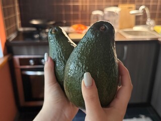 Female hands hold two big avocados against a blurred kitchen background. Fresh tropical fruit...