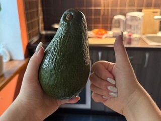 Female hands hold a giant avocado while showing a thumbs up gesture against a blurred kitchen...