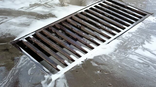 Heavy metal drain grate on a wet industrial concrete floor. White foam and water flow into the dark rusty drainage system.