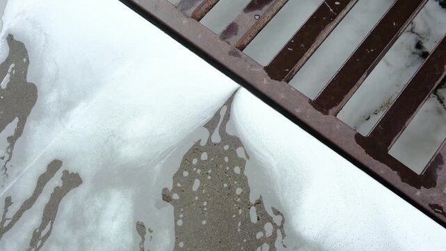 Thick white soapy foam washes over a textured gray concrete surface, flowing into an old metal storm drain grate.