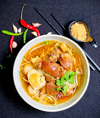 Thai braised pork leg noodle soup served in a bowl with fresh herbs, boiled egg, and green onions....