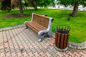 Park bench and trash can in a well-maintained green space with trees during a sunny day in a city...