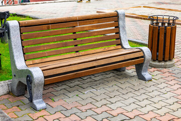 Wooden bench and trash can on a paved walkway in a public park located in a bustling city