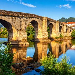 Fototapeta premium Ancient stone arch bridge spanning a calm river, reflecting the structure and surrounding landscape under a clear sky