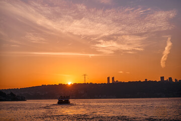 Fototapeta premium Vivid orange and pink hues fill the evening sky, casting a serene glow over silhouetted city buildings and a tranquil waterway, featuring a distant boat peacefully drifting atop the calm waters.