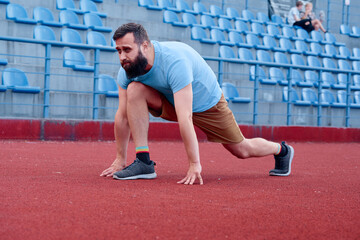 Man wearing casual athletic clothing doing stretches on a running track near stadium seats, demonstrating preparation for sports or fitness routines.