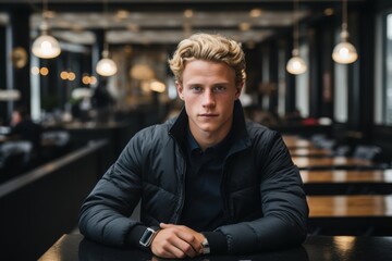A young man with blond hair sits at a table in a stylish cafe, wearing a black jacket. He gazes confidently at the camera, surrounded by ambient lighting and modern decor