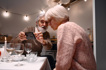 An elderly couple enjoying a serene evening together, sharing memories and photos during a cozy home dinner. The warm lighting and glassware enhance the intimate and cheerful ambiance.