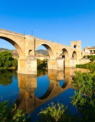 Fototapeta premium Ancient stone bridge reflected in calm river water under a clear blue sky