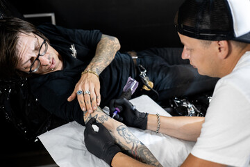 Tattoo artist working on a client’s arm in a studio, with framed tattoo designs on the wall. Concept of body art, lifestyle, creativity, and modern tattoo culture.