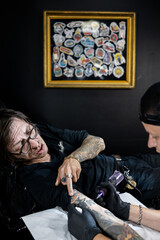Tattoo artist working on a client’s arm in a studio, with framed tattoo designs on the wall. Concept of body art, lifestyle, creativity, and modern tattoo culture.