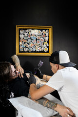 Tattoo artist working on a client’s arm in a studio, with framed tattoo designs on the wall. Concept of body art, lifestyle, creativity, and modern tattoo culture.