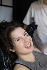 Smiling young woman getting a face tattoo in a modern studio, expressing confidence and joy during...