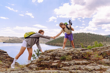 Two hikers helping each other while navigating rocky terrain near a lake during a sunny day