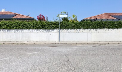 White concrete fence with hedge on top at roadside. Blank sign on pole. Roofs with solar panels and...