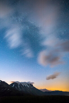 Night sky with stars and clouds over mountains. Castelluccio di Norcia, Pian Grande, Umbria, Marche, Italy. The Milky Way is visible above the landscape.