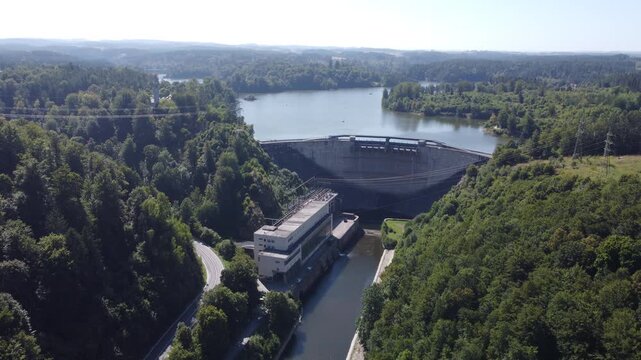Aerial view of Ottenstein power plant in Austria with a large concrete dam, lush green forest, reservoir lake, and hydroelectric station surrounded by nature and power lines under clear skies.