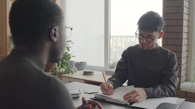 Over-the-shoulder shot of Hispanic HR manager sitting in front of young Black man aking questions about previous job and making notes