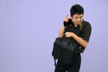 man holding a briefcase with batik outfit against a purple background, looking hesitant