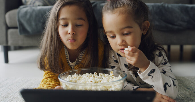 Child, siblings or watching with popcorn on tablet in home for online entertainment or subscription. People, girl or sisters streaming with technology for movie, snack or cartoon together on floor - Powered by Adobe