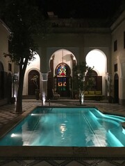 Elegant Moroccan riad courtyard at night with a glowing turquoise pool, arched architecture, trees,...