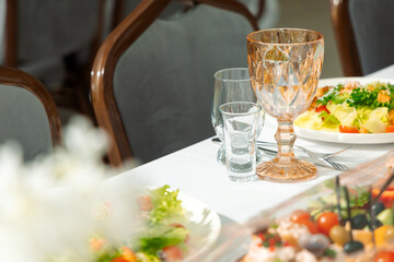 Banquet table with exquisite table setting: wine glasses, shot glasses, salad plates, fresh vegetables, greens, elegant chairs, cozy atmosphere.