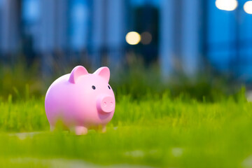 A small pink piggy bank stands on green grass, against the backdrop of blurred lights and an...
