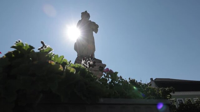 A stone statue of a robed figure stands tall in Obernberg Marktplatz, silhouetted against a bright sun in a clear blue sky. The statue is framed by vibrant red flowers and lush green foliage on a