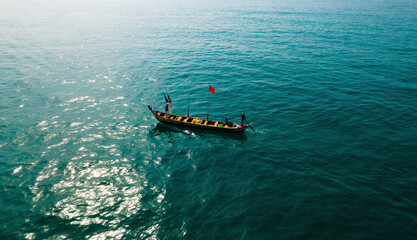 Aerial view of a traditional wooden boat cuts through the shimmering turquoise water, a vibrant red flag fluttering against the vast expanse of the sea, Location data omitted.