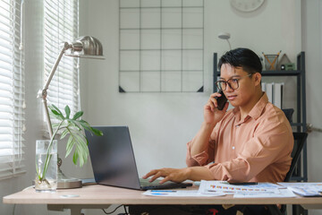 Asian freelancer talking on phone and working on laptop at home office