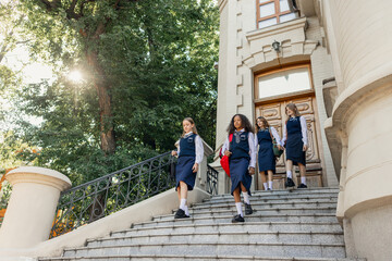 Fototapeta premium Group of schoolgirls in uniform walking down stone stairs with confidence. Concept of schoolwear fashion, education promotion, teen friendship and campus lifestyle.