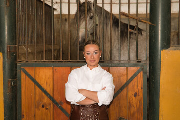 Young, beautiful, blonde woman in white shirt and leather chaps, with her arms crossed, leaning on...