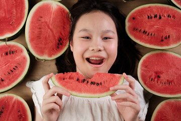 Portrait of a cheerful Asian young teenage girl eating many half pieces of fresh red watermelons,...