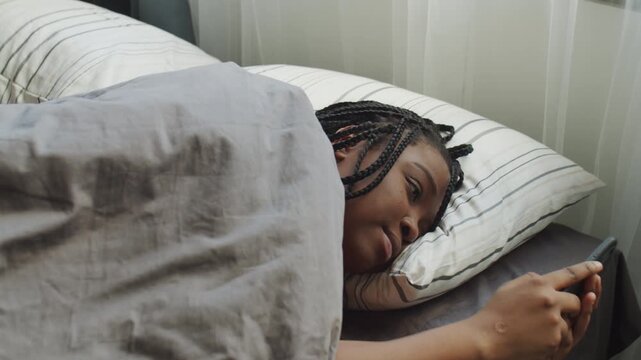 High angle sho of young Black woman waking up on weekend morning taking smartphone and checking messenger