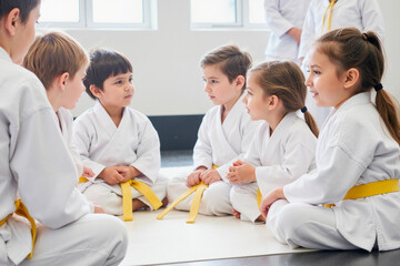 Group of diverse children sitting in circle wearing martial arts uniforms and yellow belts, engaging in conversation during karate class, focused expressions on young faces