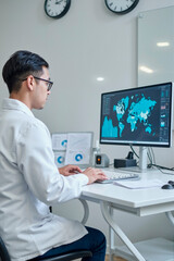 Asian young adult man sitting at desk analyzing global data on large computer monitor in modern office, wearing lab coat and working with statistical charts and world map graphics