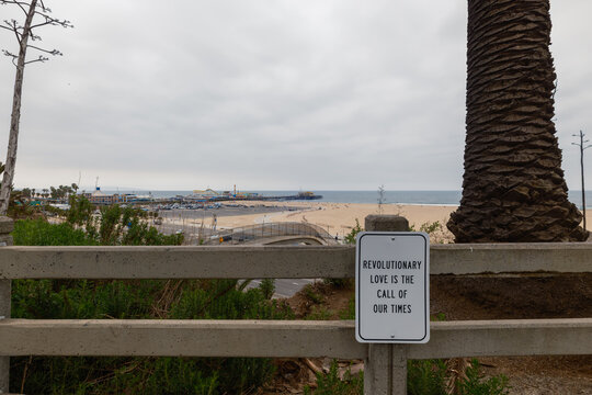View of a sign with a revolutionary message stands in contrast to the serene beach and pier, reflecting the call of our times, Santa Monica, California, United States.