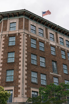 View of a stately brick building with symmetrical windows and decorative stone trim, crowned by a waving American flag against a cloudy sky, Los Angeles, California, United States.