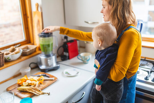 Woman in the kitchen preparing healthy smoothie and holding her baby son