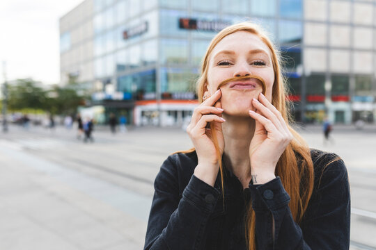 Portrait of redheaded young woman making mustache with her hair, Berlin, Germany
