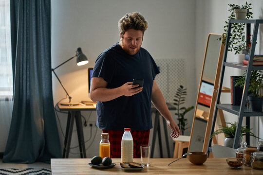 Horizontal medium shot of young Caucasian man checking e-mail on smartphone before having breakfast