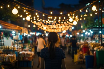 Fototapeta premium A woman walks through a bustling outdoor market filled with stalls and shoppers, warmly lit by countless string lights creating a lively nighttime atmosphere