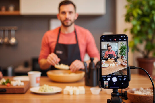 Caucasian young adult man preparing food in kitchen while being recorded on smartphone mounted on tripod, focusing on cooking process with ingredients visible on table