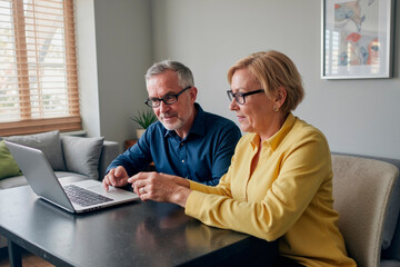 Caucasian middle aged man and Caucasian middle aged woman sitting together using laptop computer, both wearing glasses and focusing on screen, collaborating on project at home