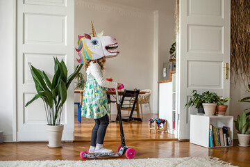 Girl with unicorn mask riding push scooter at home